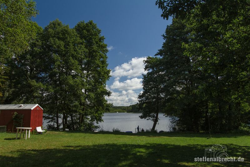 Foto Campingplatz: eine Wiese mit Bäumen am See, darauf ein rotes Holzhäuschen